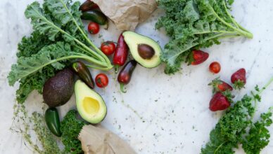 Top view of fresh avocados, kale, tomatoes, and peppers arranged on a marble surface.