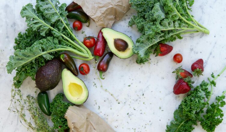 Top view of fresh avocados, kale, tomatoes, and peppers arranged on a marble surface.